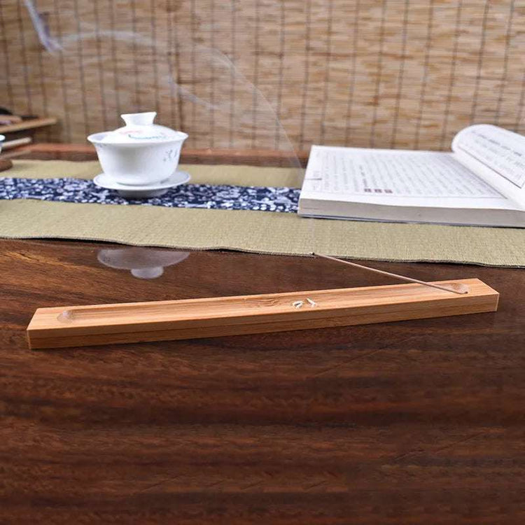 Wooden incense stick holder with smoke rising, placed on a wooden table with tea set and open book in the background.